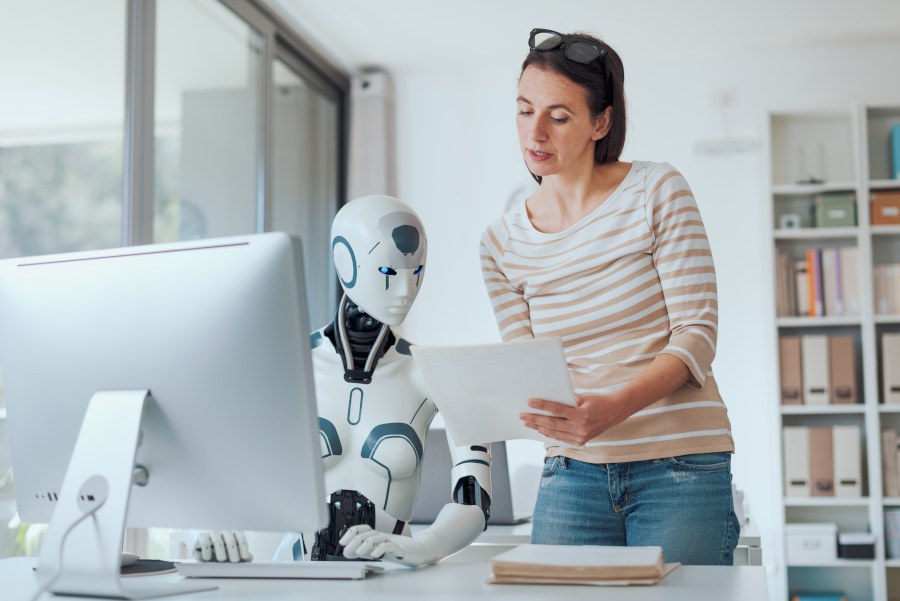 Ein KI-Roboter sitzt vor einem PC, eine Frau steht daneben und erklärt ihm etwas.