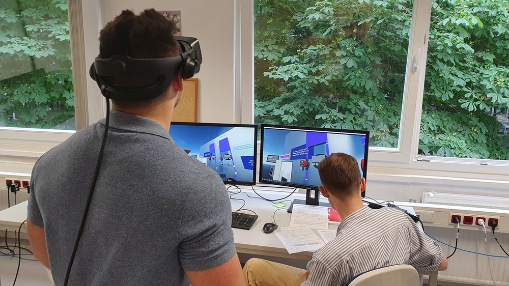 Zwei Studierende sind von hinten zu sehen, einer steht und hat eine VR-Brille auf, der andere sitzt an einem Tisch, vor ihm zwei Bildschirme, die eine VR-Umgebung zeigen.. 