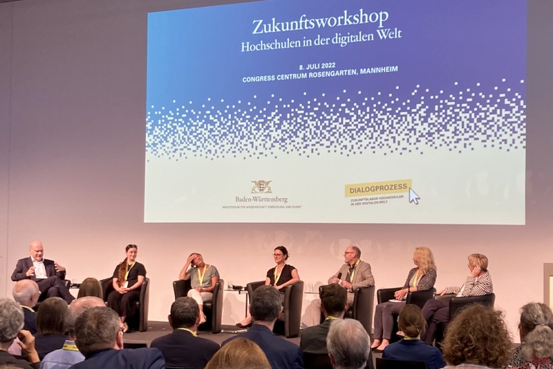 Sieben Podiumsgäste sitzen in einem Halbkreis auf der Bühne. Im Hintergrund ist ein Bild an die Wand projiziert, auf dem steht "Zukunftsworkshop Hochschulen in der Digitalen Welt"