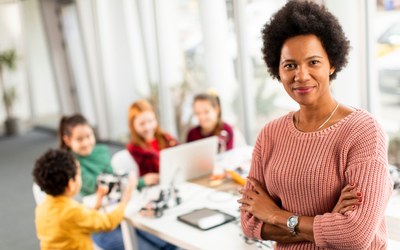 Lehrerin steht im Vordergrund und schaut in die Kamera, hinter ihr sind vier Kinder zu sehen, die an einem Tisch sitzen und in einen Laptop schauen.